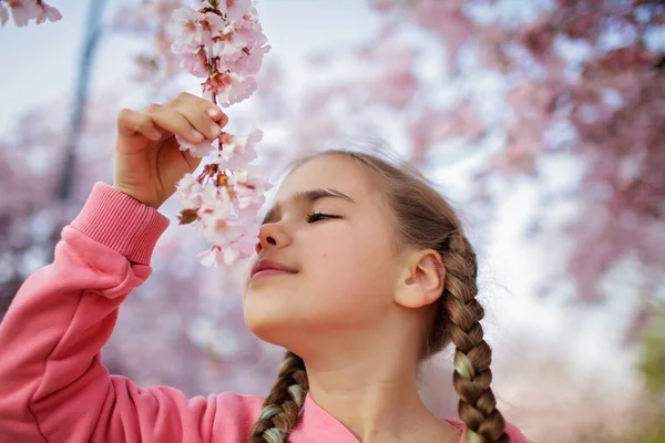 Preteen girl smells a blooming branch of sakura while walking in blossom garden. Happy spring vibes — Stock Photo, Image