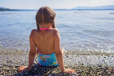 Tatlı kız dinleniyor ve taşların üzerinde oturuyor Cenevre Gölü 'nün çakıl taşları üzerinde güneş ışığı altında mavi dağların üzerinde hayal kuruyor ve mutlu, yaz tatili, seyahat ve yaz tatili, yaşam tarzı, arka plan