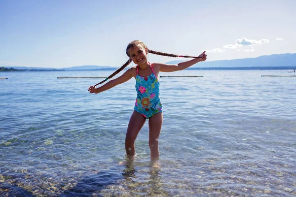 Happy girl with two pigtails has fun on pebble beach on Geneva Lake over blue mountains in sun light, lots of fun and happiness, summer vacations, travel and summertime, lifestyle