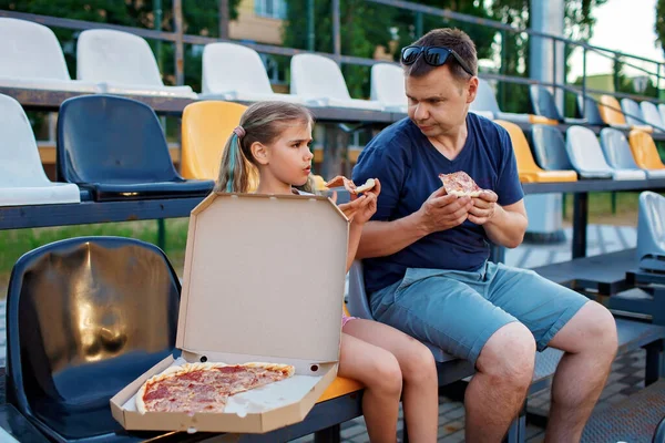 Father with daughter, soccer fans, watching football match, eating pizza and cheering for local team at stadium, real emotions, sports event and fan supporting, outdoor lifestyle, street food