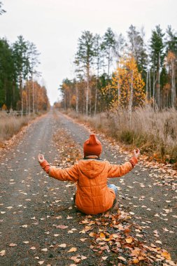 Ten years girl in orange coat sits in yoga pose on the road in fall forest. Dry grass and golden trees. Autumn vibes, outdoor lifestyle. Deep in thoughts, daydreaming and mental health