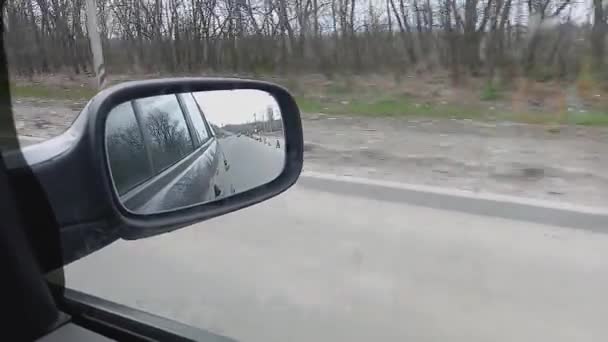 A dark gray car drives down a road in Ukraine during martial law. The side mirror shows anti-tank hedgehogs, tires and traffic cones — Stock Video