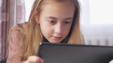 Genç bir kız elinde tabletle bir odada bir yatakta yatıyor. Tablette oynayan bir kızın tam yüzü, beyaz gün. Yüksek kalite 4k görüntü