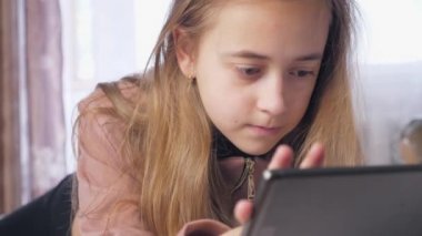 Genç bir kız elinde tabletle bir odada bir yatakta yatıyor. Tablette oynayan bir kızın tam yüzü, beyaz gün. Yüksek kalite 4k görüntü