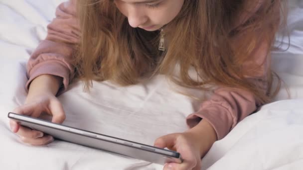 10代の少女がタブレットを持った部屋のベッドの上に横たわっている タブレットで遊んでいる少女の完全な顔 白い日 高品質4K映像 — ストック動画