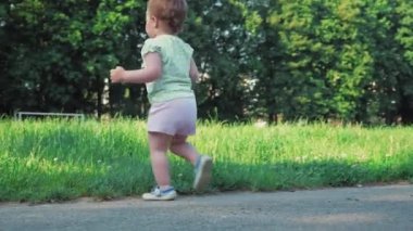 Küçük bir kız yeşil çimlere doğru koşar. Tişörtlü ve şortlu küçük bir kız parkta yürüyor. Çocuk uzun ağaçların arka planında çimenlerde koşuyor. Yüksek kalite 4k görüntü