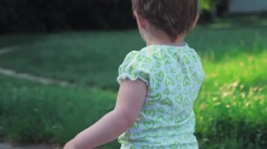 Küçük bir kız yeşil çimlere doğru koşar. Tişörtlü ve şortlu küçük bir kız parkta yürüyor. Çocuk uzun ağaçların arka planında çimenlerde koşuyor. Yüksek kalite 4k görüntü