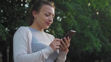Güzel bir kız sokakta duruyor ve elinde bir telefon tutuyor. Genç bir kadın tarayıcıda zaman geçirir. Kız doğada olduğu için sosyal medyada geziniyor. Yüksek kalite 4K görüntü