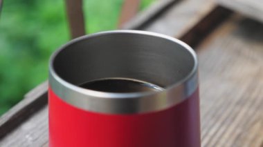 Beyaz süt, içine kahve dökülen kırmızı bir termo bardağa dökülür. Sütlü kahve yapma süreci. Kırmızı bir kupa, yeşil ağaçlara bakan bir pencerenin kenarındaki ahşap pencere eşiğinde duruyor. Yüksek