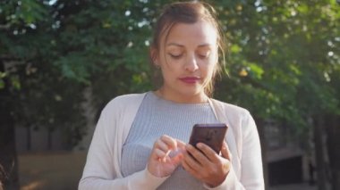 Güzel bir kız sokakta duruyor ve elinde bir telefon tutuyor. Genç bir kadın tarayıcıda zaman geçirir. Kız doğada olduğu için sosyal medyada geziniyor. Yüksek kalite 4K görüntü