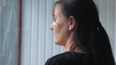 Siyah saçlı genç bir kadın balkondaki pencerenin yanında duruyor ve kırmızı bardaktan kahve içiyor. Güzel bir kız sırtı verandada kameraya dönük duruyor ve manzaranın tadını çıkarıyor.