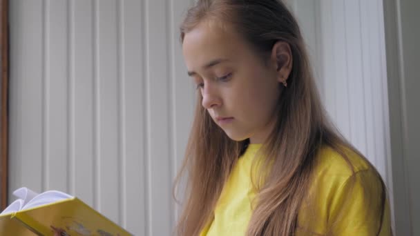 一个穿着黄色T恤衫的可爱的金发少女正在看一本黄色的书 她站在阳台上 窗外阳光灿烂 参天大树清晰可见 — 图库视频影像