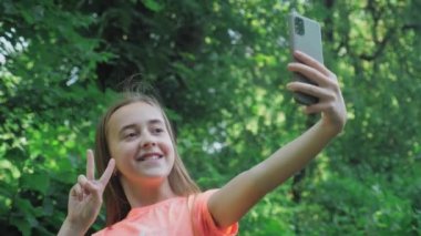 Uzun saçlı güzel bir genç kız elinde bir telefon tutuyor ve doğanın arka planına karşı selfie çekiyor. Telefon kamerasına poz veren sevimli küçük bir kız. Yüksek kalite 4k görüntü