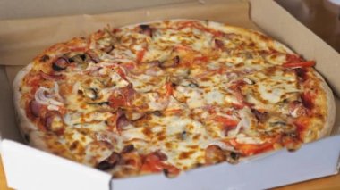 Hot appetizing pizza with cheese, Bavarian sausages, tomatoes, onions and mushrooms lies in a cardboard box on the table. High quality 4k footage