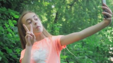 Uzun saçlı güzel bir genç kız elinde bir telefon tutuyor ve doğanın arka planına karşı selfie çekiyor. Telefon kamerasına poz veren sevimli küçük bir kız. Yüksek kalite 4k görüntü