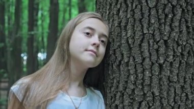 Sevimli genç kız ormanda bir ağaç gövdesine yaslanmış duruyor. Uzun bir ağacın yanında düşünceli bir kız. Yüksek kalite 4k görüntü