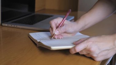 Bir kadın tükenmez kalemle deftere yazıyor. Defter, dizüstü bilgisayarın yanındaki masanın üzerinde. Kadınların elleri kapalı olsun. Yüksek kalite 4k görüntü