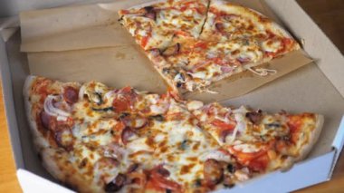 Hot appetizing pizza with cheese, Bavarian sausages, tomatoes, onions and mushrooms lies in a cardboard box on the table. Appetizing pizza lies in a box, a few pieces of pizza are missing. The opened