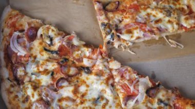 Hot appetizing pizza with cheese, Bavarian sausages, tomatoes, onions and mushrooms lies in a cardboard box on the table. Appetizing pizza lies in a box, a few pieces of pizza are missing. The opened