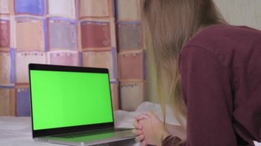 A cute teenage girl is lying on the bed, in front of her is a laptop with a green screen. A teenager is alone in her room looking at a laptop screen. High quality 4k footage