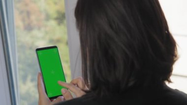A girl with short dark hair stands with her back to the camera and holds a phone with a green screen. A woman makes purchases on the site using the phone. The girl stands in front of the window in the