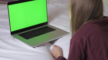 A cute teenage girl is lying on the bed, in front of her is a laptop with a green screen that has gone out. Teenager alone in his room, looking at a laptop screen. High quality 4k footage