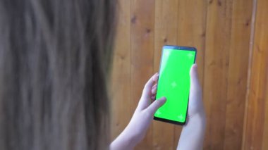 A teenage girl holds a phone with a green screen in her hands while standing on a balcony. Caucasian girl surfs the phone instead of doing her homework. High quality 4k footage