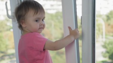 Küçük bir kız, yüksek katlı bir binanın penceresinin yanındaki pencere pervazında duruyor ve elleriyle yarı açık pencereye tutunuyor. Çocuk apartmanın penceresinden caddeye bakıyor.