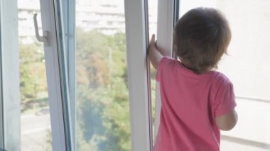 Küçük bir kız, yüksek katlı bir binanın penceresinin yanındaki pencere pervazında duruyor ve elleriyle yarı açık pencereye tutunuyor. Çocuk apartmanın penceresinden caddeye bakıyor.