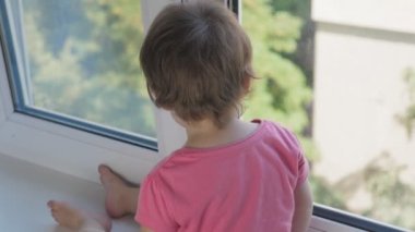 Küçük bir çocuk pencere pervazında oturur ve pencereden şehre bakar. İhmalkârlık ve sorumsuzluk kavramı. Bebek yüksek katlı bir binanın penceresinden dışarı bakıyor. Yüksek kalite 4k