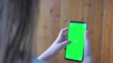 A teenage girl holds a phone with a green screen in her hands while standing on a balcony. Caucasian girl surfs the phone instead of doing her homework. High quality 4k footage