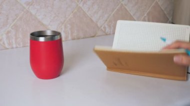 Kız mutfak masasında oturup sıcak kahve içerken bir not defterine not alıyor. Kadınların elleri kapalı olsun. Bir kadın sıcak bir içecek içer ve bir günlüğe fikirler yazar. 18.09.2022 Ukrayna