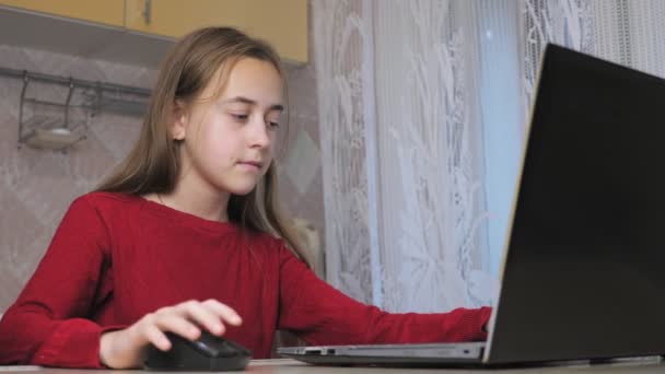 赤いセーターを着た10代の女の子が台所の椅子に座っている間 ラップトップでビデオゲームをプレイします 子供は宿題をするのではなくノートパソコンで遊ぶ 高品質4K映像 — ストック動画