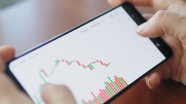 Kadın akıllı telefondaki dijital bir borsada fiyat tablosunu kontrol ediyor. Elinde cep telefonu olan eller, borsa verilerini kontrol ediyor. Dokunmatik ekran aygıtındaki borsa grafiğine yakınlaştırılıyor, kaydırılıyor