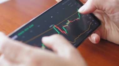 Kadın akıllı telefondaki dijital bir borsada fiyat tablosunu kontrol ediyor. Elinde cep telefonu olan eller, borsa verilerini kontrol ediyor. Dokunmatik ekran aygıtındaki borsa grafiğine yakınlaştırılıyor, kaydırılıyor