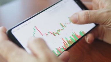 Kadın akıllı telefondaki dijital bir borsada fiyat tablosunu kontrol ediyor. Elinde cep telefonu olan eller, borsa verilerini kontrol ediyor. Dokunmatik ekran aygıtındaki borsa grafiğine yakınlaştırılıyor, kaydırılıyor