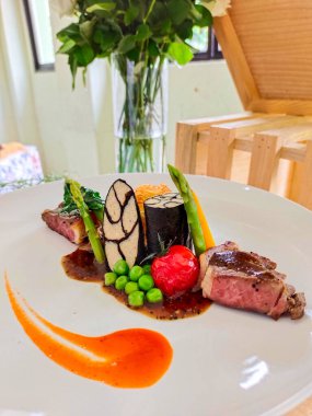 Kuşkonmaz, havuç, domates ve bezelye ile kaplı beyaz bir tabakta servis edilen sığır filetosu ve balık rolladı.