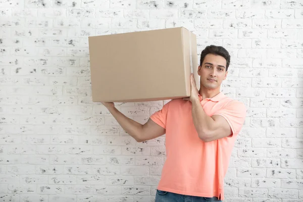 Handsome man carrying huge and heavy box in front of white brick wall background