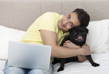 Yatakta oturmuş gülümseyen mutlu bir adam sevimli siyah pug cinsini kucaklıyor ve dizüstü bilgisayarıyla çalışıyor. Arkadaşlık kavramı