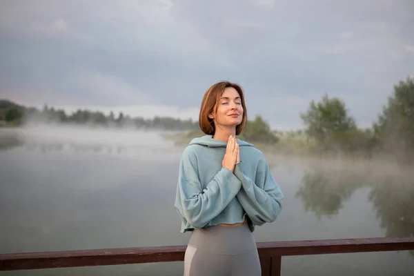 Portrait of young and beautiful woman praying and meditating alone on the sunrise at the coast of the foggy lake. Sensitivity to nature, spiritual and emotional concept.
