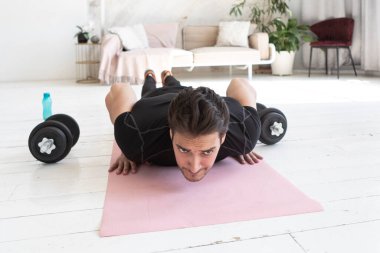 Genç sporcu evde eğitim görüyor. Spor giyim bölümündeki yakışıklı adam halterlerle antrenman yaptıktan sonra yorgun ve oturma odasındaki spor paspasına uzanmış.