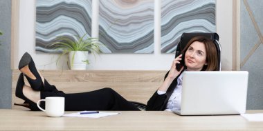 Kendine güvenen ve çekici bir iş kadını. Bacakları masanın üstünde, cep telefonuyla konuşuyor ve modern ofiste dizüstü bilgisayarla çalışıyor.