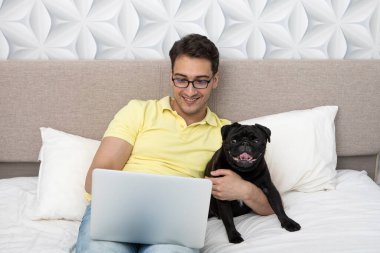 Yatakta oturmuş gülümseyen mutlu bir adam sevimli siyah pug cinsini kucaklıyor ve dizüstü bilgisayarıyla çalışıyor. Arkadaşlık kavramı