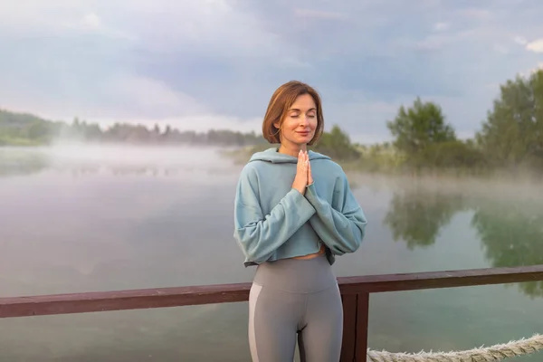 Portrait of young and beautiful woman praying and meditating alone on the sunrise at the coast of the foggy lake. Sensitivity to nature, spiritual and emotional concept.