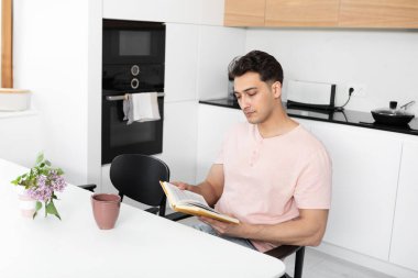 Genç yakışıklı adam kitap okuyor ve mutfakta çay içiyor.