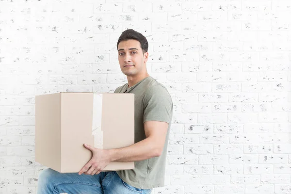 Handsome man carrying huge and heavy box in front of white brick wall background
