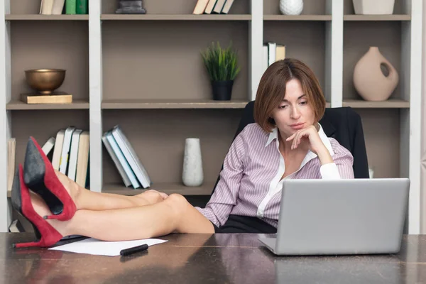 Young confident and attractive business woman with legs on desk and working on laptop in the modern office