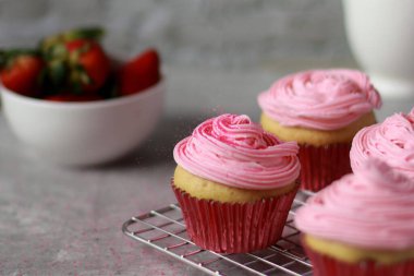 Kekler üzerine pembe tereyağlı krema süslemesi ile süslenmiş kırmızı kablar, gri mermer masa ve beyaz taş duvar