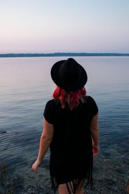 woman standing at the beach, pink hair and black hat, back view