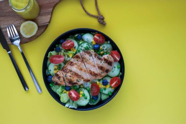 grilled chicken salad on black plate with fork and knife, honey mustard sauce, yellow background with copy space